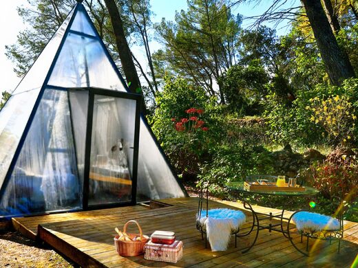 Cabane tipyramide vitrée sur terrasse bois, chaises cosy et verdure, séjour insolite au cœur de la nature. Auberge de la Tuilière