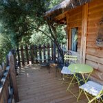 Terrasse bois avec chaises jaunes, table verte, près d'une cabane perchée entourée de nature. Auberge de la Tuilière