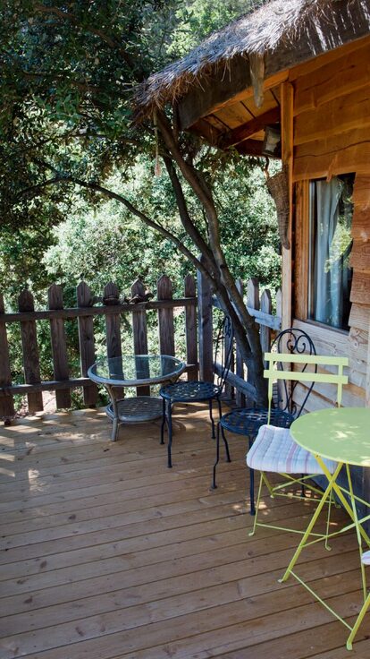 Terrasse bois avec chaises jaunes, table verte, près d'une cabane perchée entourée de nature. Auberge de la Tuilière