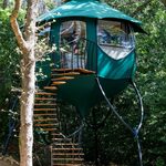 Cabane perchée moderne, sur pilotis noirs, escalier spiral et grandes fenêtres, au cœur d'un écrin verdoyant. Auberge de la Tuilière