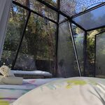 Intérieur d'une tente transparente nichée en nature, lit douillet, vue sur arbres verdoyants et lumière douce. Auberge de la Tuilière