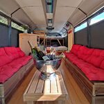 Bus aménagé cosy, banquettes bois, coussins rouges ; table d'accueil, lit surélevé, coin intime. Auberge de la Tuilière